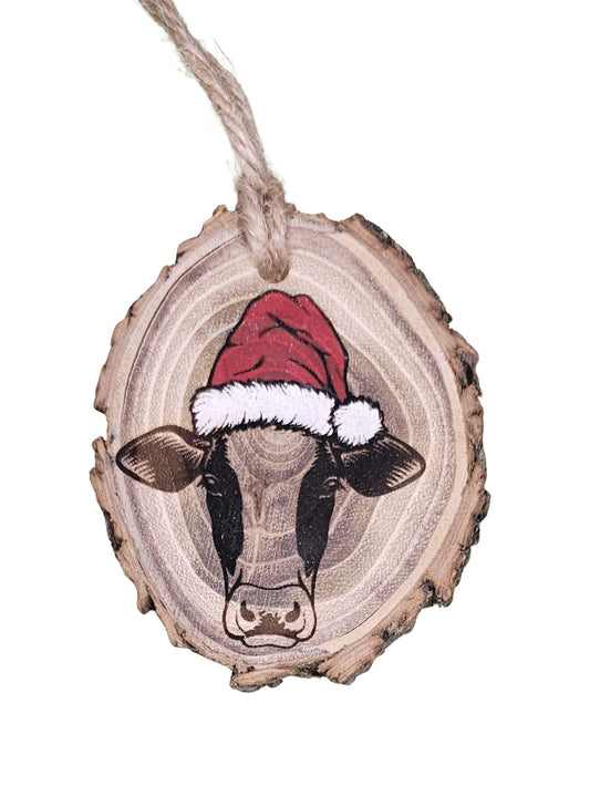A wood burned Christmas tree ornament depicting a cow with a Santa hat with a laser engraved hand drawn design, suspended by a twine string.
