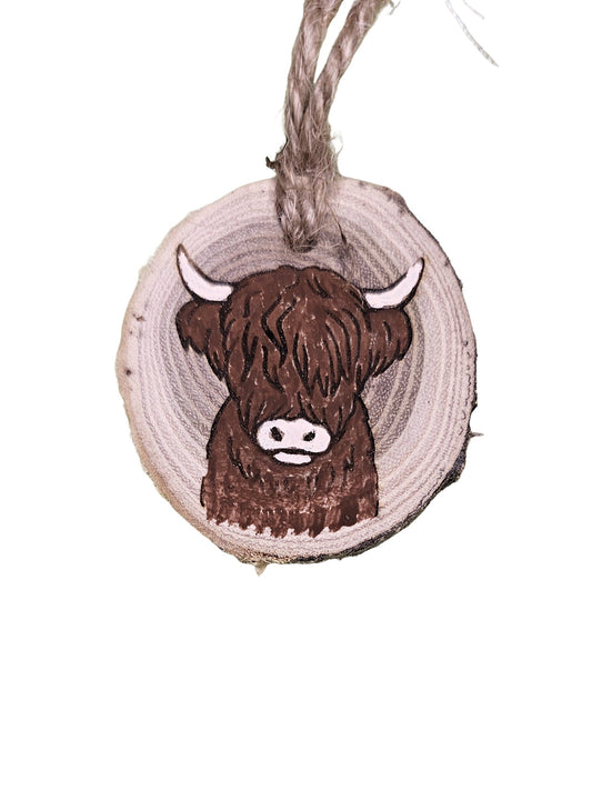 A wood burned Christmas tree ornament depicting a highland cow with a laser engraved hand drawn design, suspended by a twine string.
